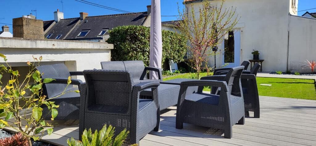 An outdoor seating area with wicker furniture on a wooden deck, surrounded by greenery and a building in the background.