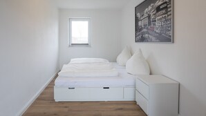 Ein schlichter Schlafzimmerbereich mit Bett, Nachttisch, Fenster und einem Stadtlandschaftsbild.