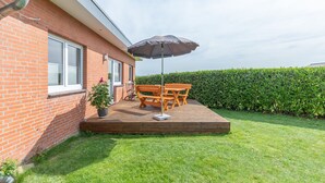 Eine Terrasse mit Holzmöbeln und einem Tisch unter einem Sonnenschirm, angrenzend an ein Ziegelgebäude mit einem Fenster und einem Heckenstreifen.