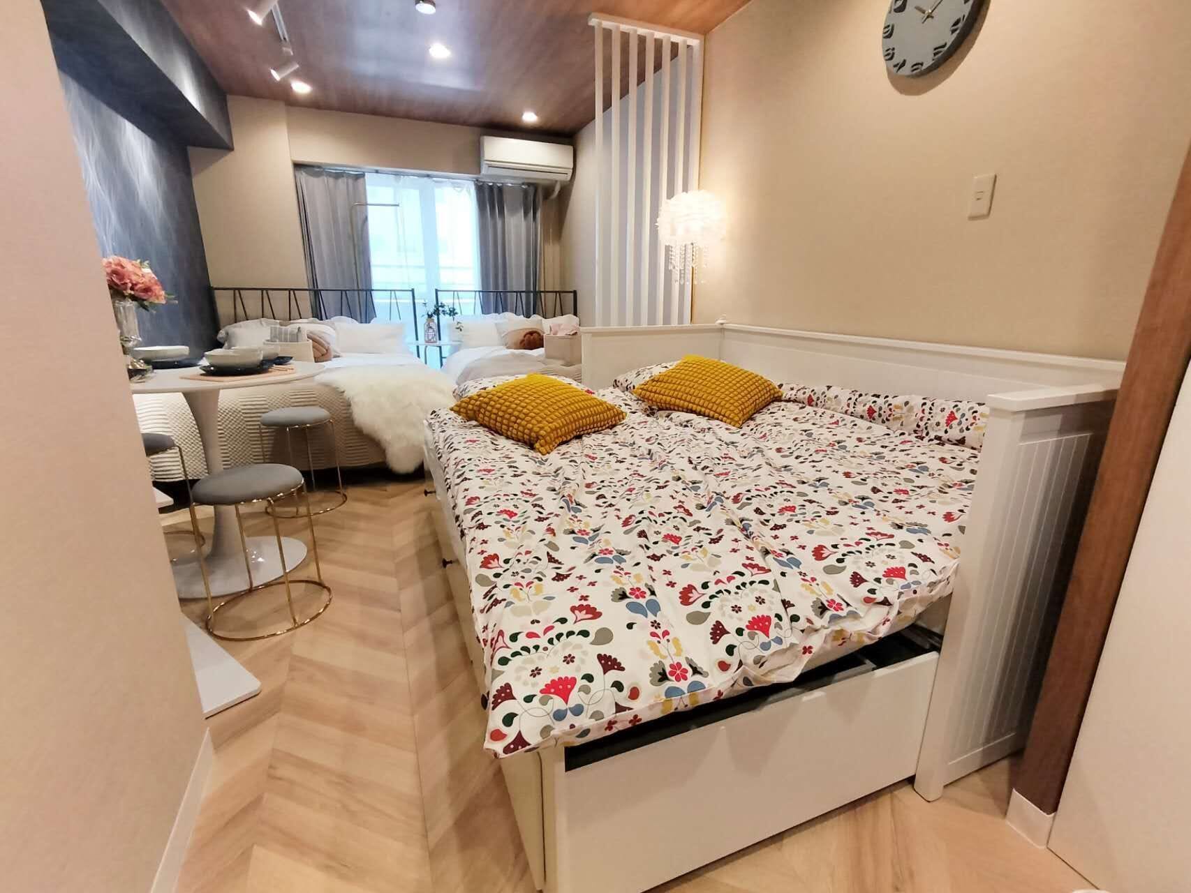 Ein kleines Zimmer mit einem Bett, einem kleinen Tisch und einem Stuhl. Es gibt ein Fenster mit Vorhängen, eine Wanduhr und eine Klimaanlage.