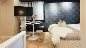 A modern hotel room with a wooden ceiling, a black patterned wall, a round dining table with chairs, a bed with a white furry throw, and a flat-screen TV mounted on the wall. - 4 min walk from the nearest station in the most prosperous town in Shinjuku City Apartment room (新宿区)