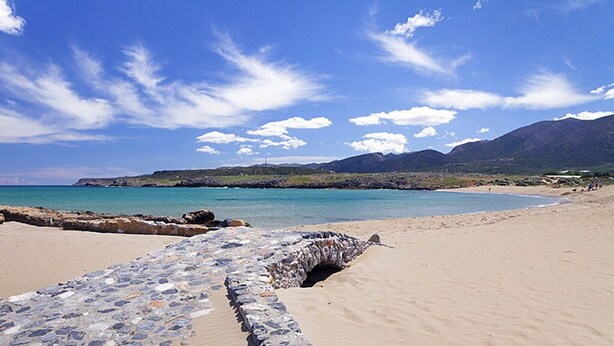 A beach with a stone wall and a clear view of the ocean and mountains in the background. - Helen Suites (Hersonissos)