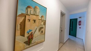 A hallway with a large painting of a building and a person. There is a green door and a fire extinguisher on the wall. - Helen Suites (Hersonissos)