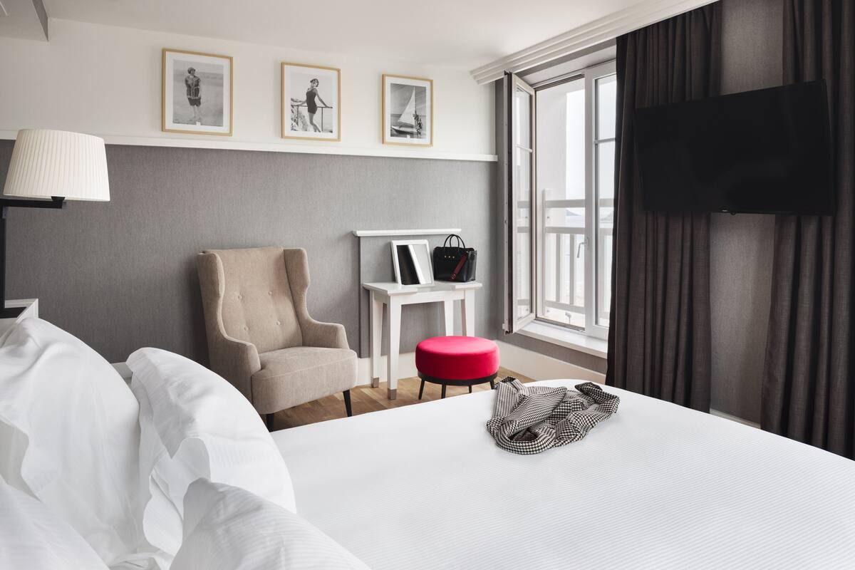 A hotel room with a bed, a chair, a small table with a handbag, and a TV mounted on the wall.