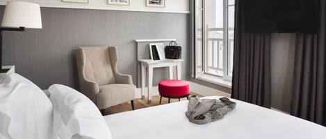A hotel room with a bed, a chair, a small table with a handbag, and a TV mounted on the wall.
