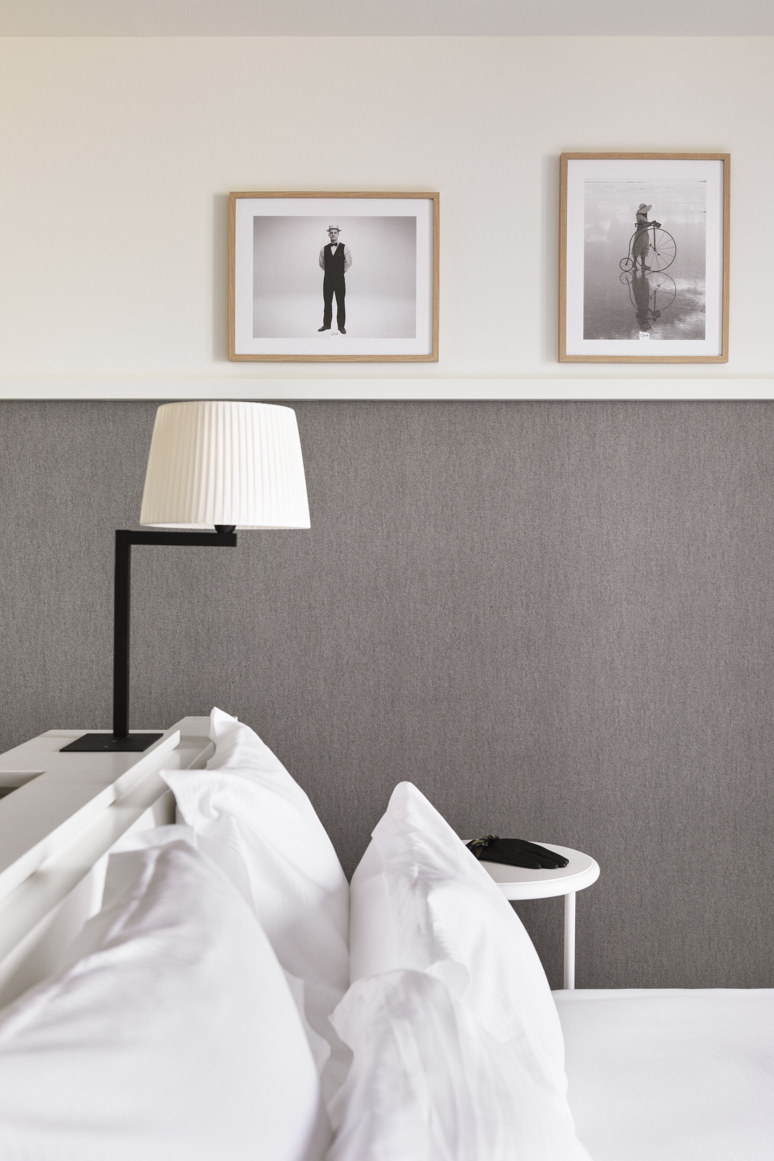 Een net opgemaakt bed met witte lakens, een zwarte vloerlamp en twee ingelijste foto's aan de muur.