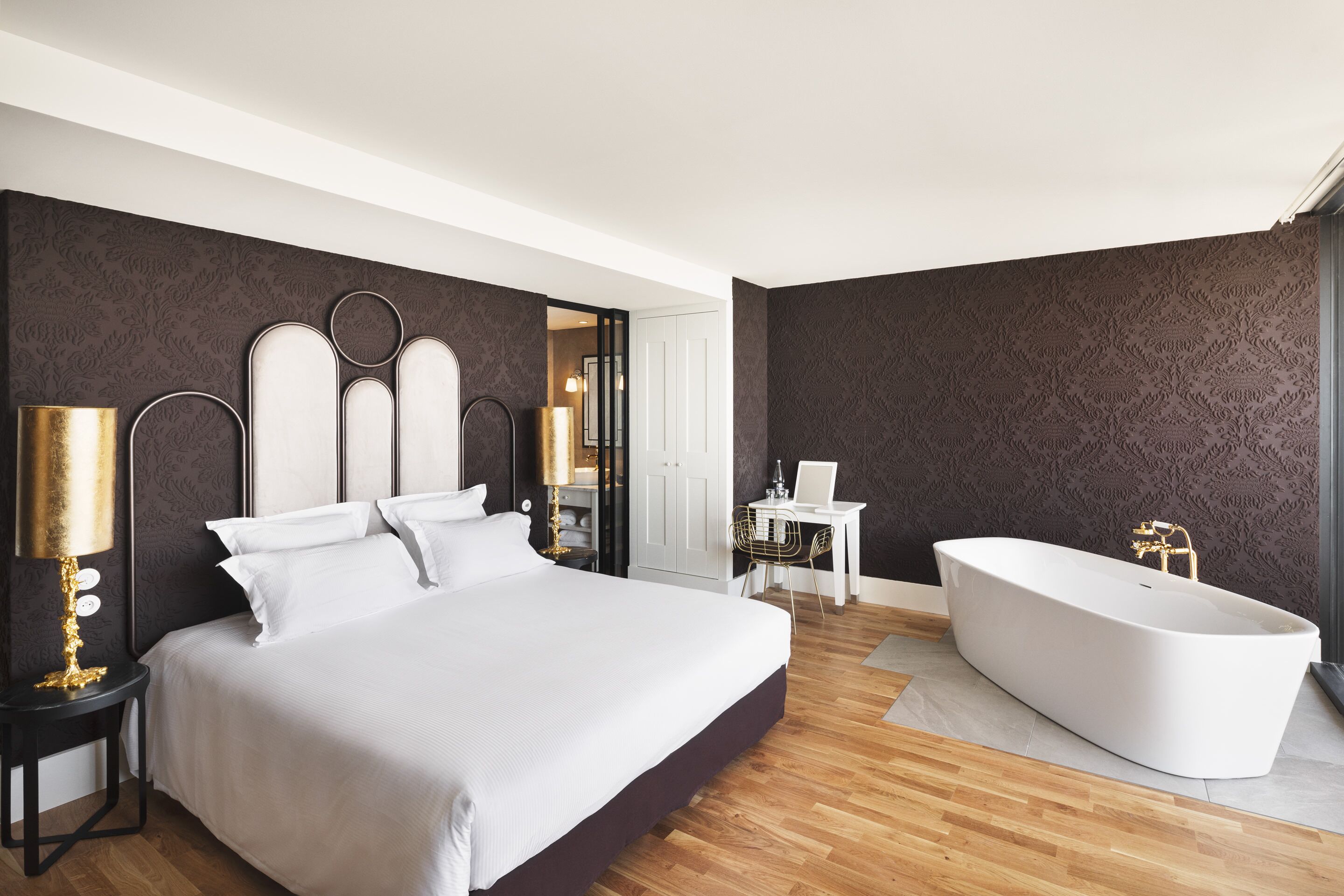 A modern bedroom with a large bed, a white bathtub, and a desk with a laptop.