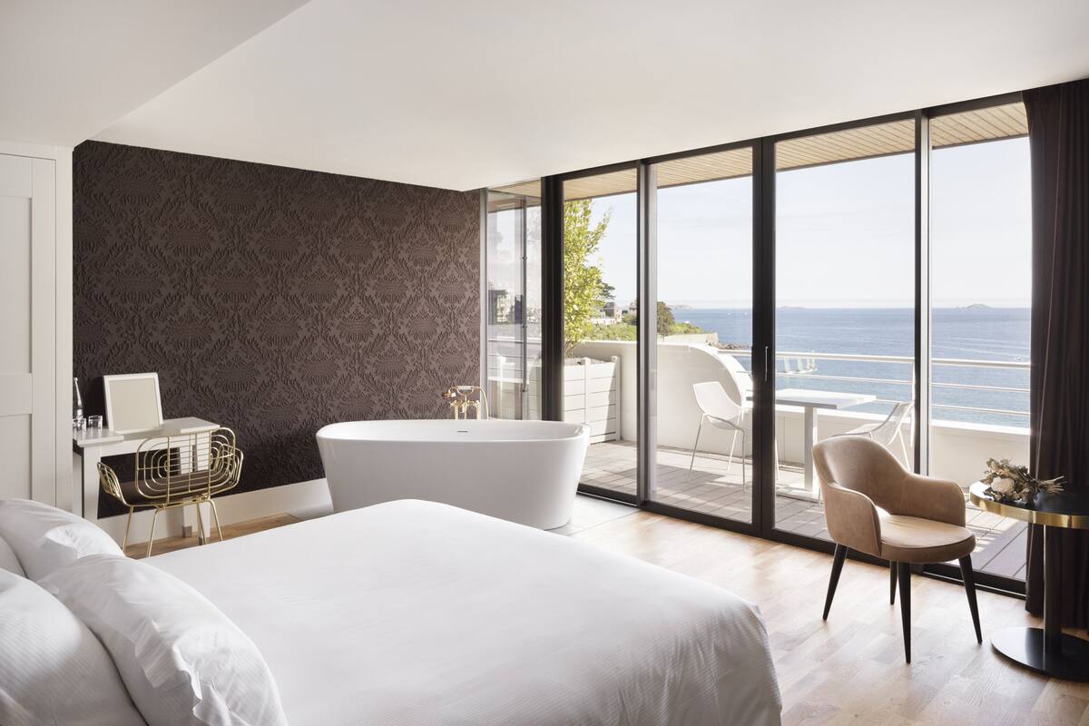 A modern hotel room with a large bed, a bathtub, a desk, and a view of the sea.