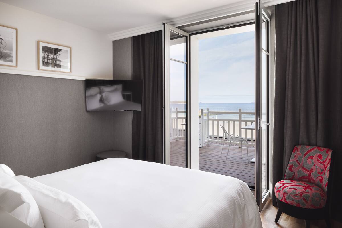 A hotel room with a bed, a TV mounted on the wall, a chair, and a balcony with a view of the sea.