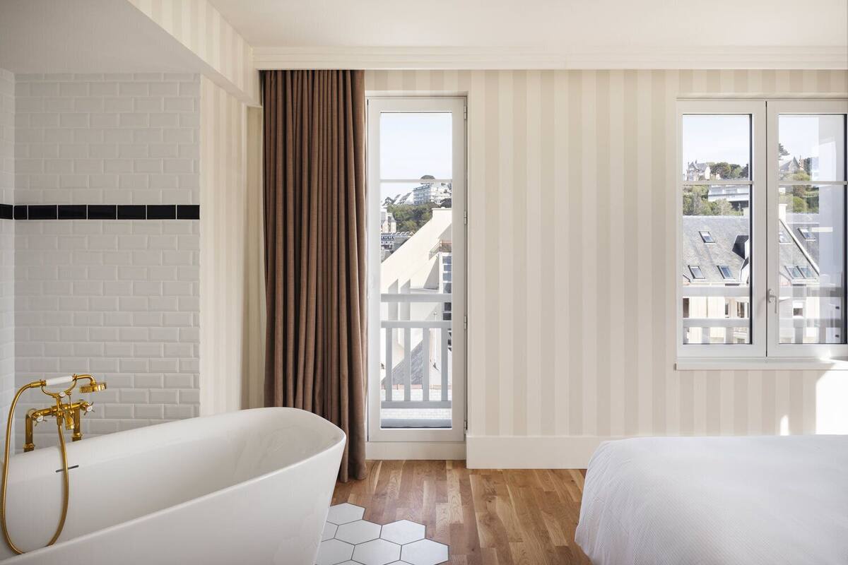 A modern bathroom with a white bathtub, a shower, and a view of a balcony with buildings.
