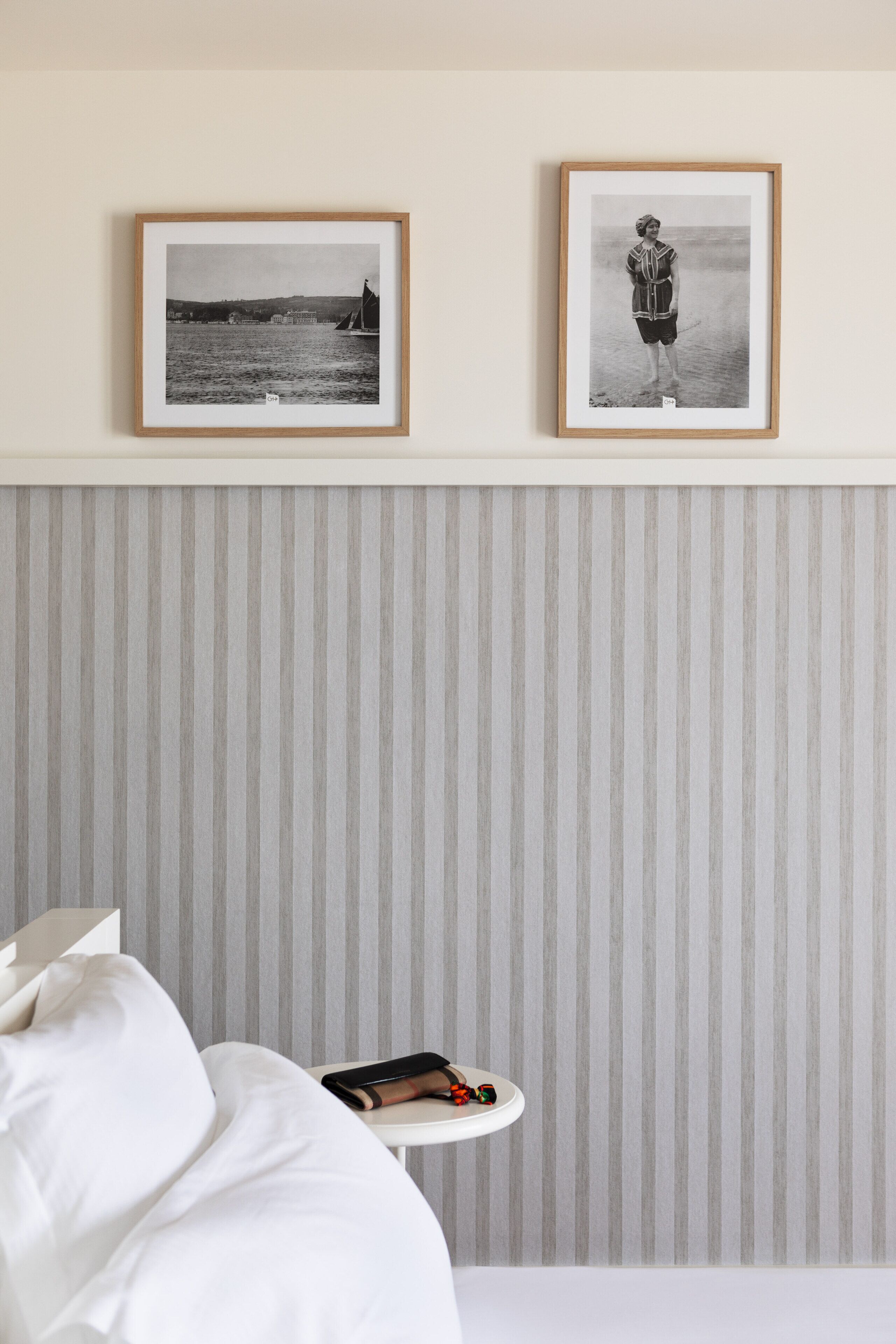 A hotel room with a bed, a bedside table with a book and a pair of scissors, and two framed black and white photographs on the wall.