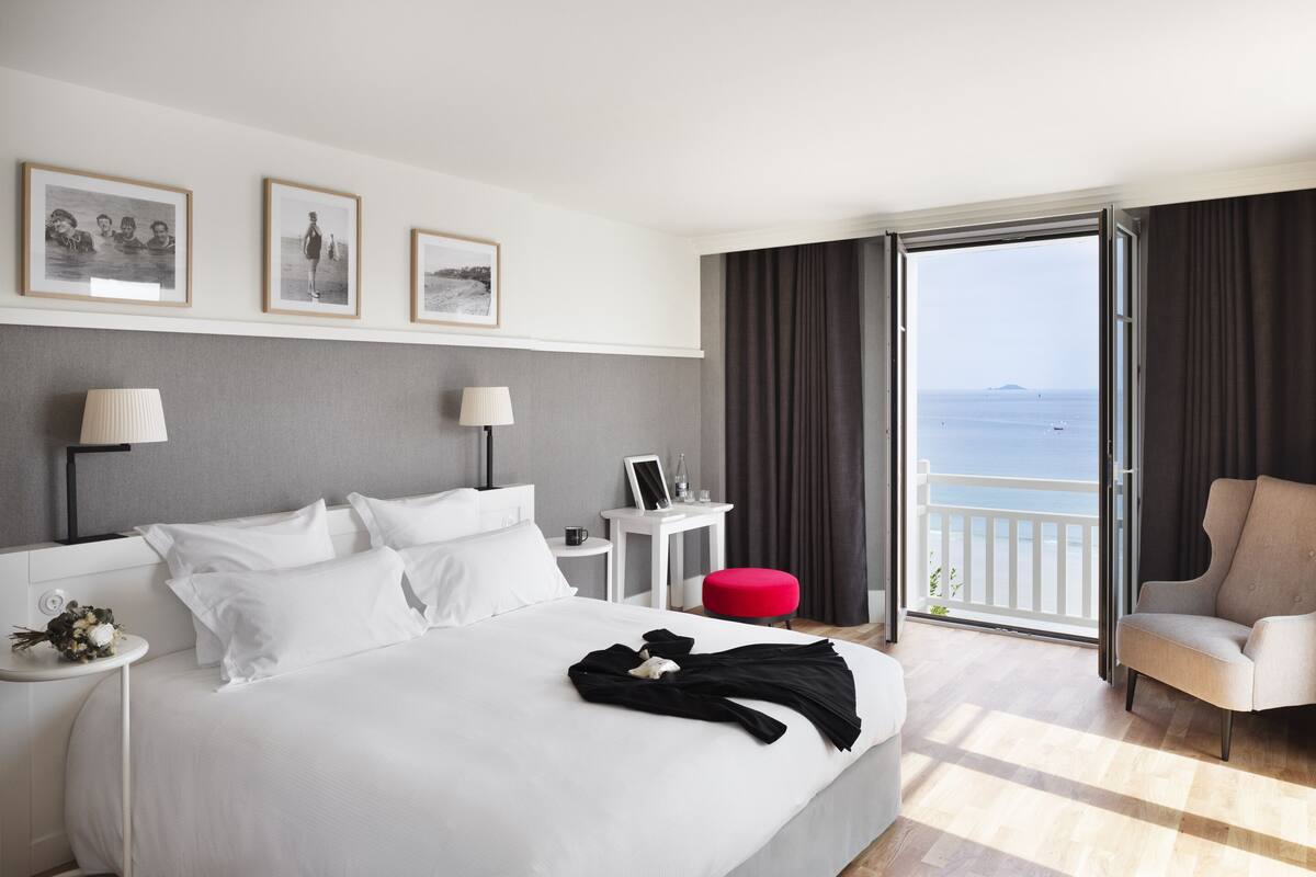 A hotel room with a large bed, a desk, a chair, and a balcony with a view of the sea.