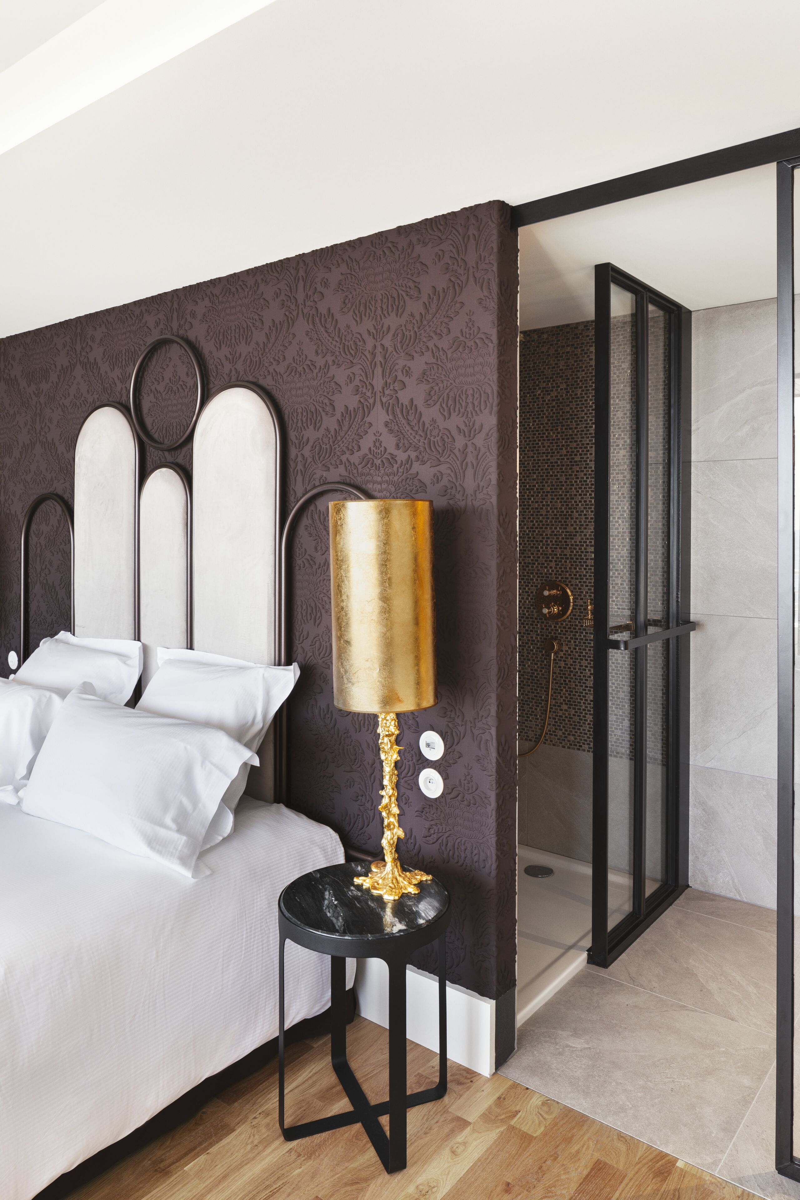 A modern hotel room with a bed, a bedside table with a lamp, a shower area, and a patterned wall.