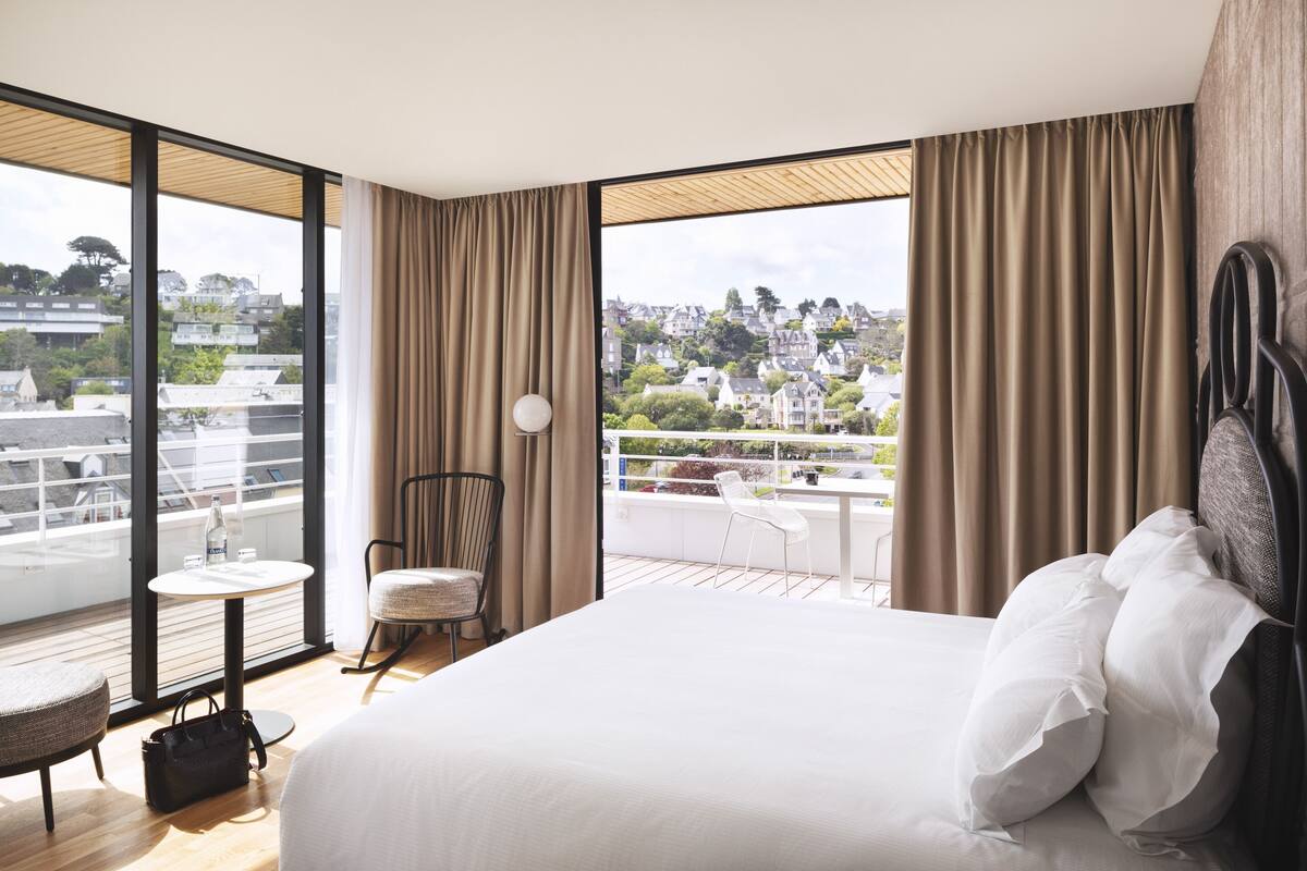 A hotel room with a large bed, a small table, a chair, and a view of a cityscape through large windows.