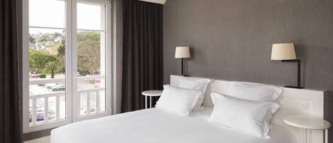 A hotel room with a large bed, two bedside tables with lamps, a view of the outside through a window, and a grey wall.