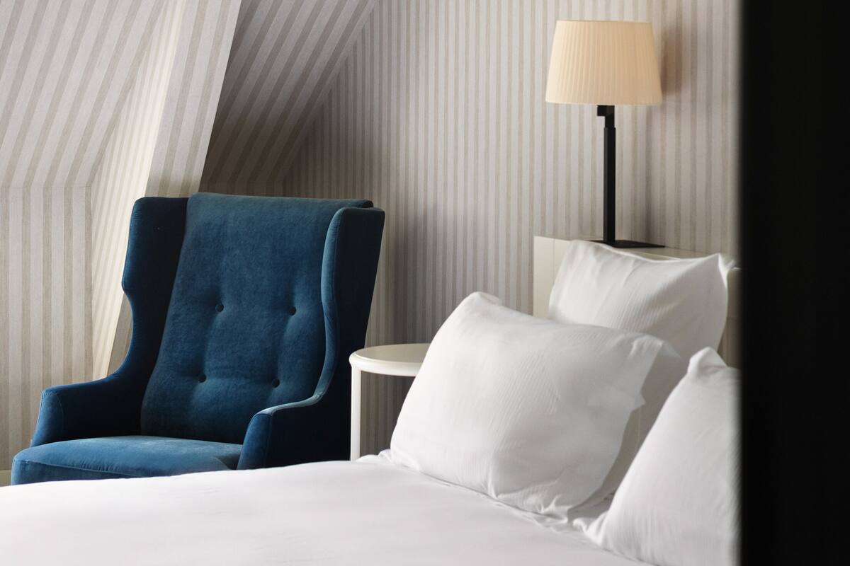 A hotel room with a bed, a blue armchair, a bedside table with a lamp, and a framed picture on the wall.