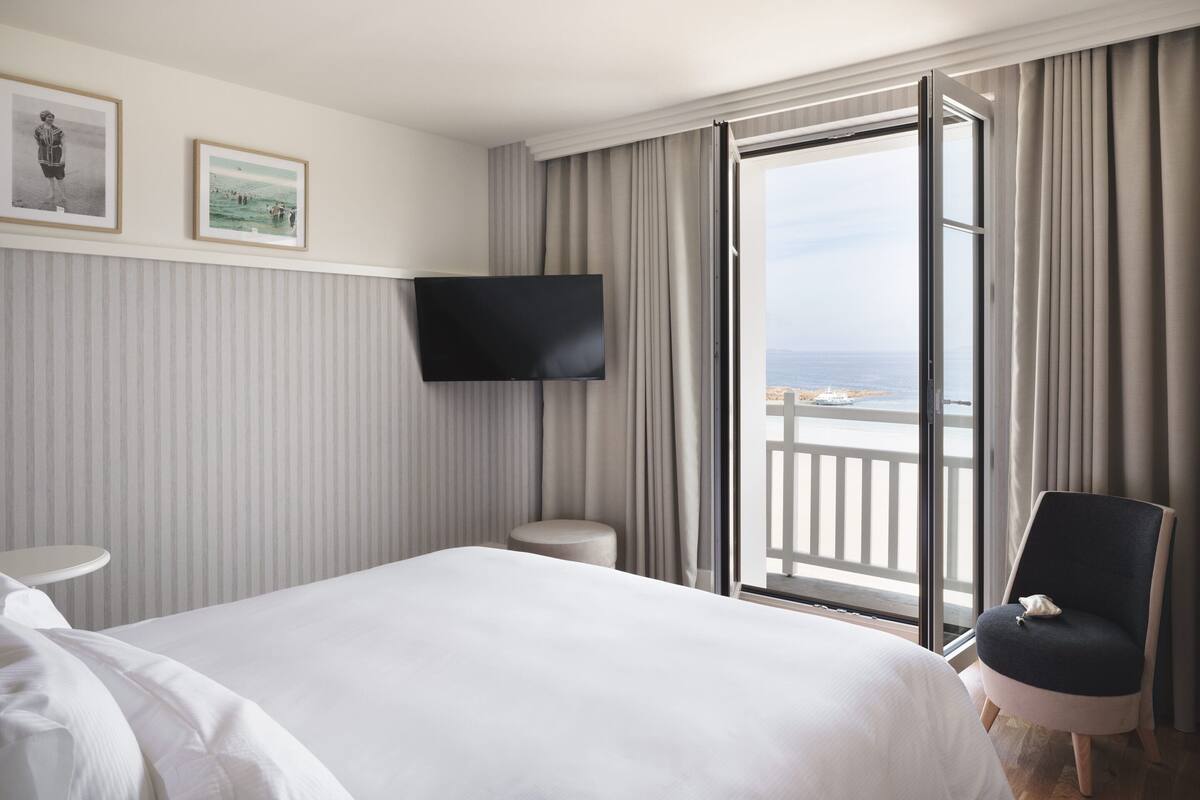 A hotel room with a bed, a TV, a chair, and a balcony with a view of the sea.