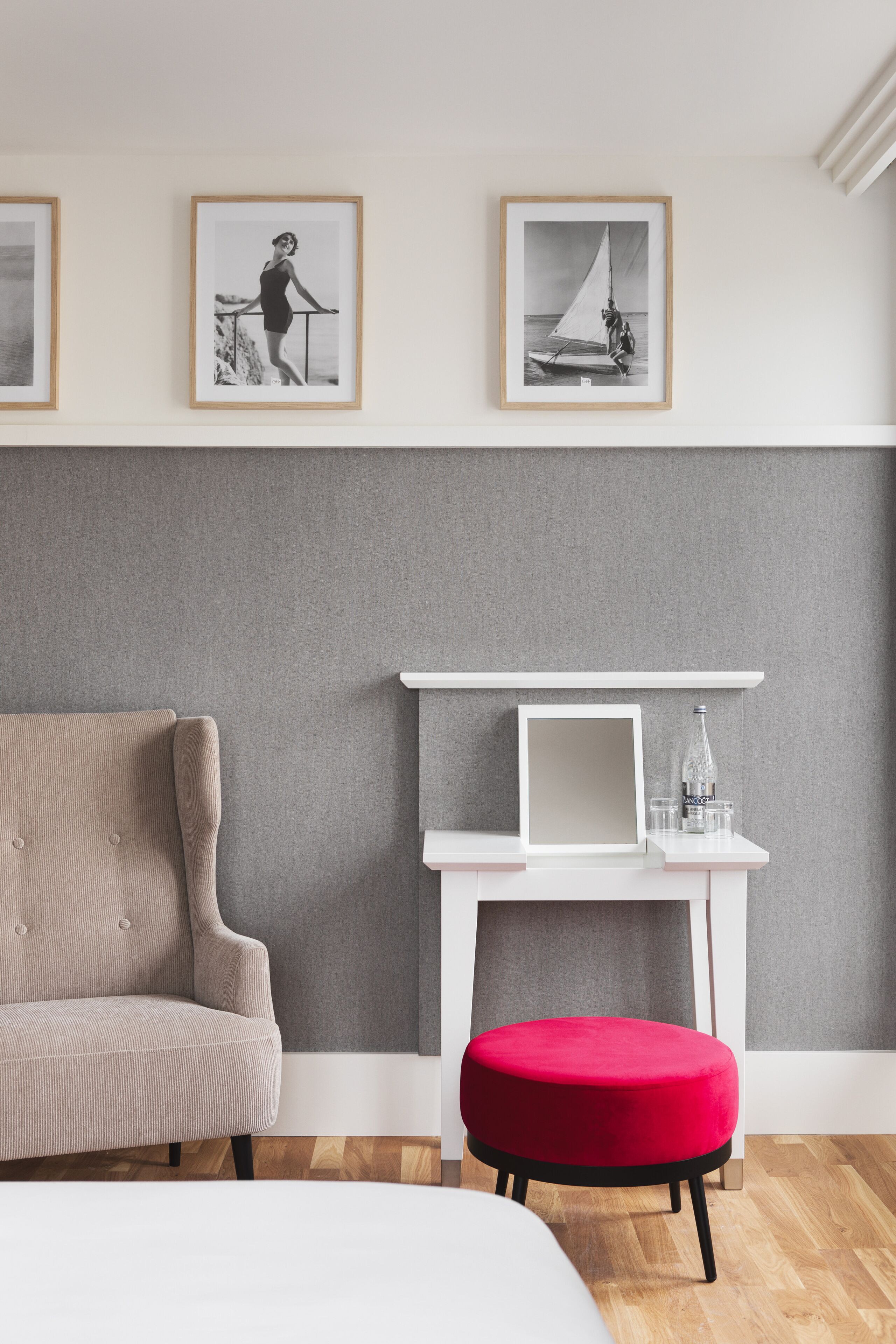 A modern room with a grey and white color scheme, featuring a sofa, a red ottoman, a white desk with a laptop and glasses, and framed artwork on the wall.