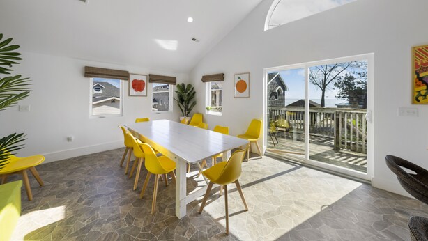 Un vaste espace dédié à la salle à manger, avec une grande table et des chaises jaunes, un sol en pierre et une vue sur la terrasse extérieure, visible à travers des portes vitrées.