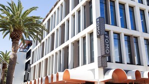Un edificio moderno con cielo despejado, que presenta un llamativo letrero 'FOMO', grandes ventanales y una palmera en primer plano.