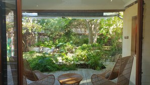 Vista a través de un ventanal de un patio con mobiliario de mimbre y un jardín.