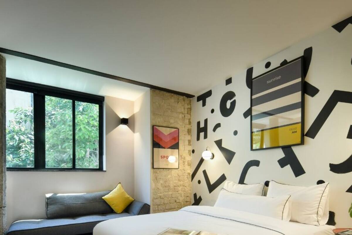 A modern bedroom with a large bed, a desk with a lamp, a window, and a wall with abstract art.