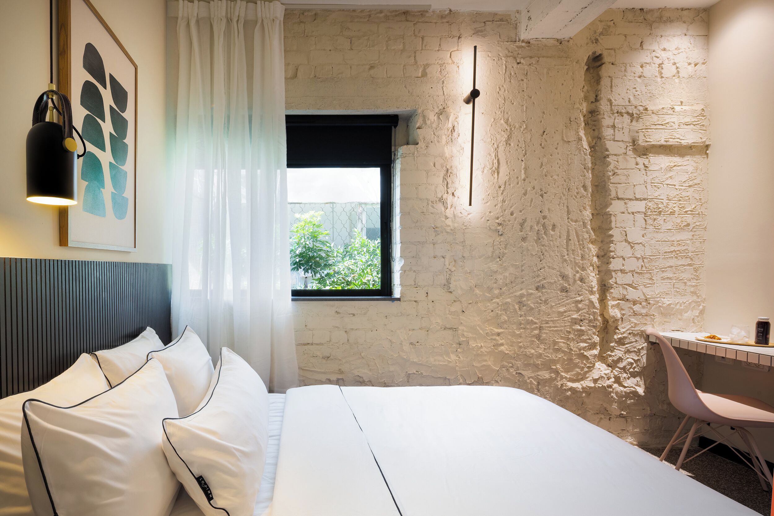 Ein Hotelzimmer mit Bett, Schreibtisch, Stuhl, Fenster mit Blick auf Grünflächen und einer Wand mit freiliegenden Ziegelsteinen.