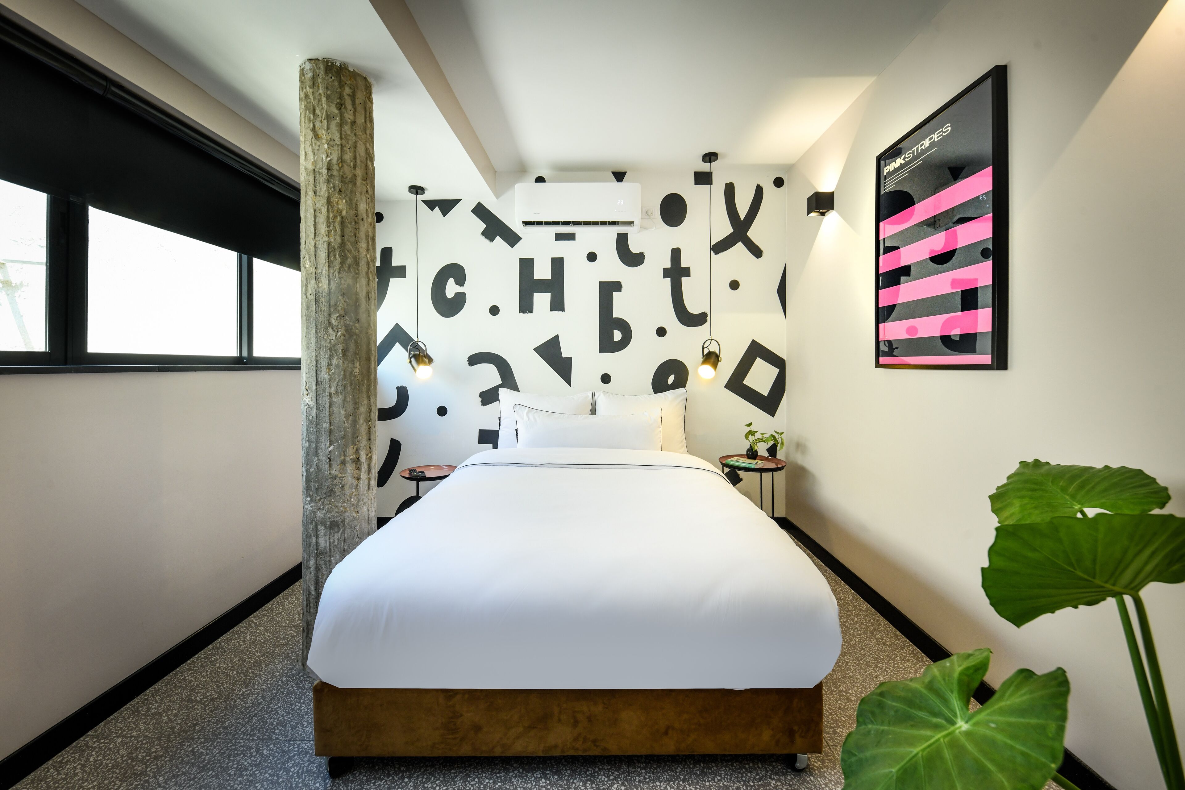 A modern hotel room with a large bed, a wall-mounted air conditioner, a small bedside table with a plant, and a poster on the wall.