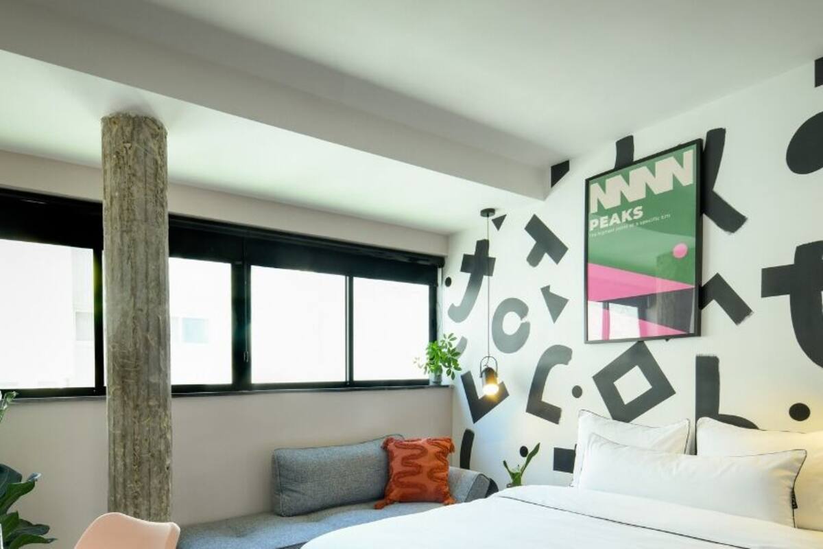 A modern bedroom with a large window, a wooden column, a bed with white bedding, and a wall with abstract black and white patterns.
