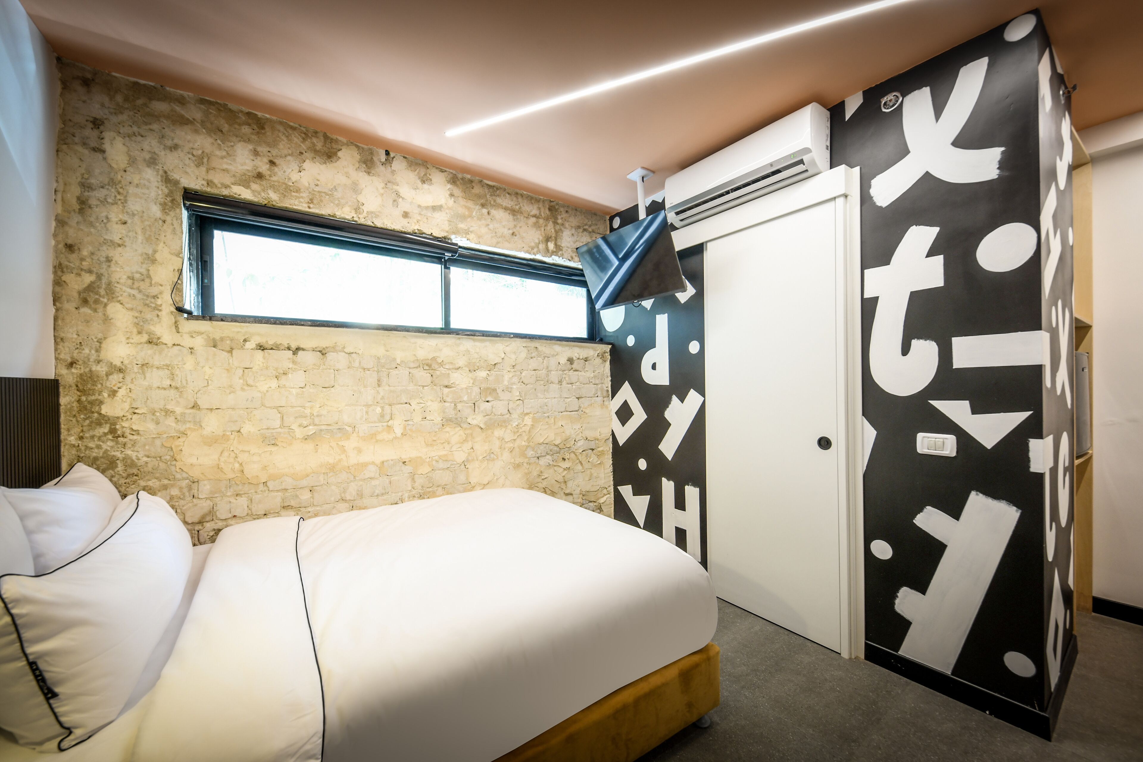 Ein Schlafzimmer mit einer Ziegelwand, einem Fenster, einem Bett mit weißer Bettwäsche und einer schwarz-weißen Tür mit Muster.