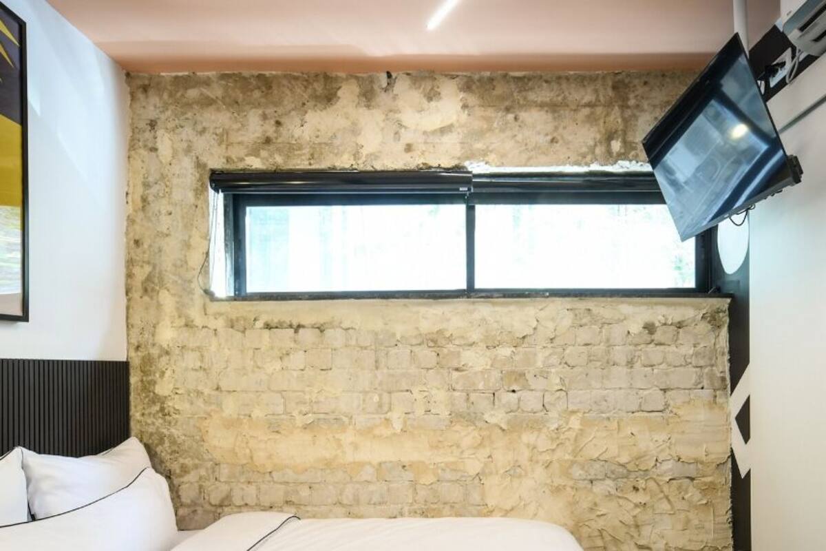 A bedroom with a brick wall, a window, a bed, a mounted TV, and a framed picture on the wall.