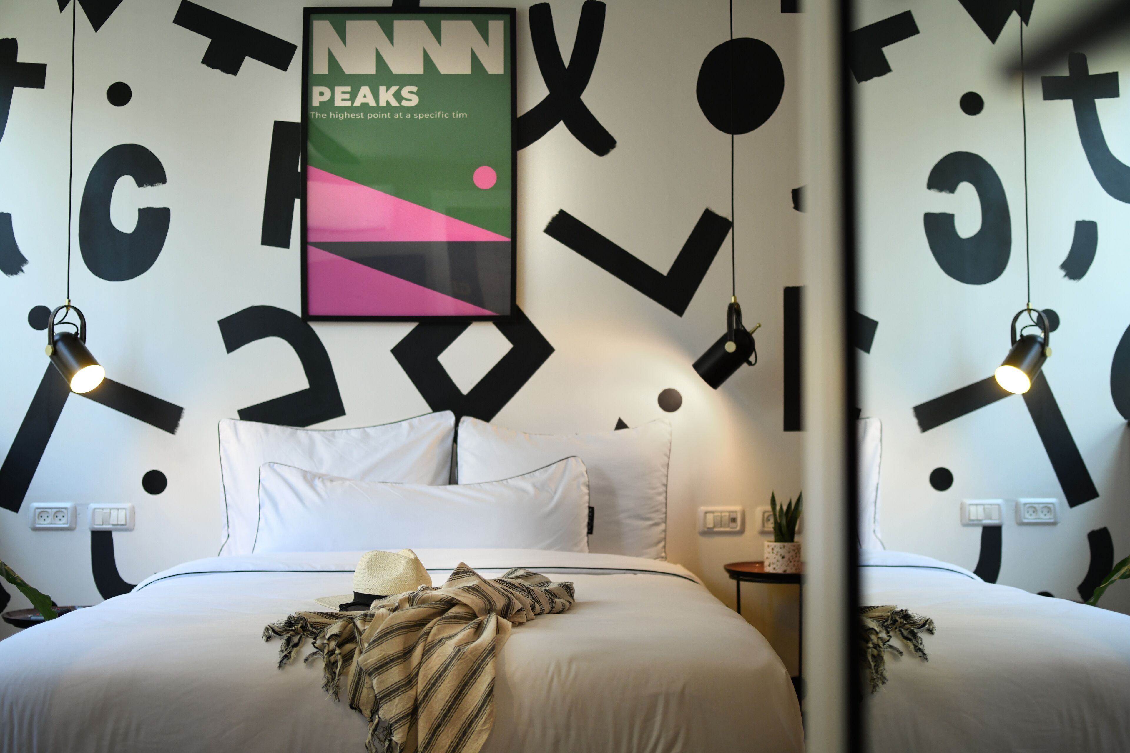Un letto rifatto con lenzuola bianche, un cuscino decorativo e una coperta a righe. Sulla parete sopra il letto è appeso un poster con la scritta “NNN PEAKS”.