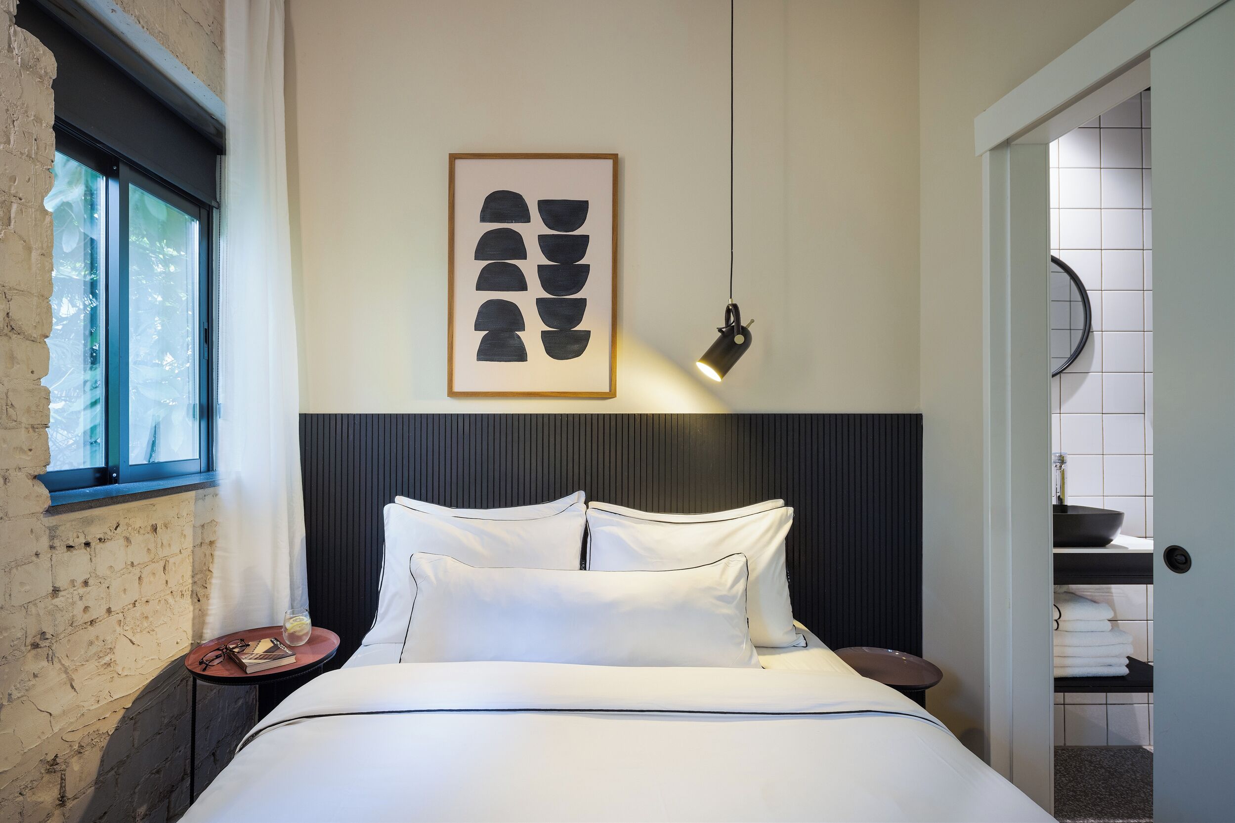 Habitación de hotel con cama, mesita de noche, una obra de arte enmarcada, una lámpara colgante y vistas al baño.