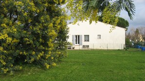 A white building with a door and windows, surrounded by a grassy yard and a tree with yellow flowers.