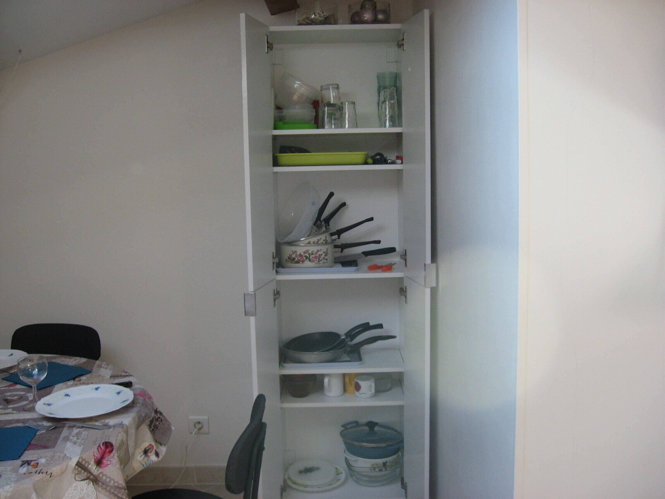 A kitchen with a white cabinet containing various kitchenware, a dining table with a floral tablecloth, and a chair.