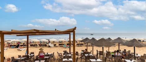 A beachside restaurant with outdoor seating, umbrellas, and a view of the ocean.