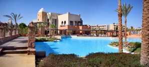 A resort with a large swimming pool, palm trees, and a building with arched windows.