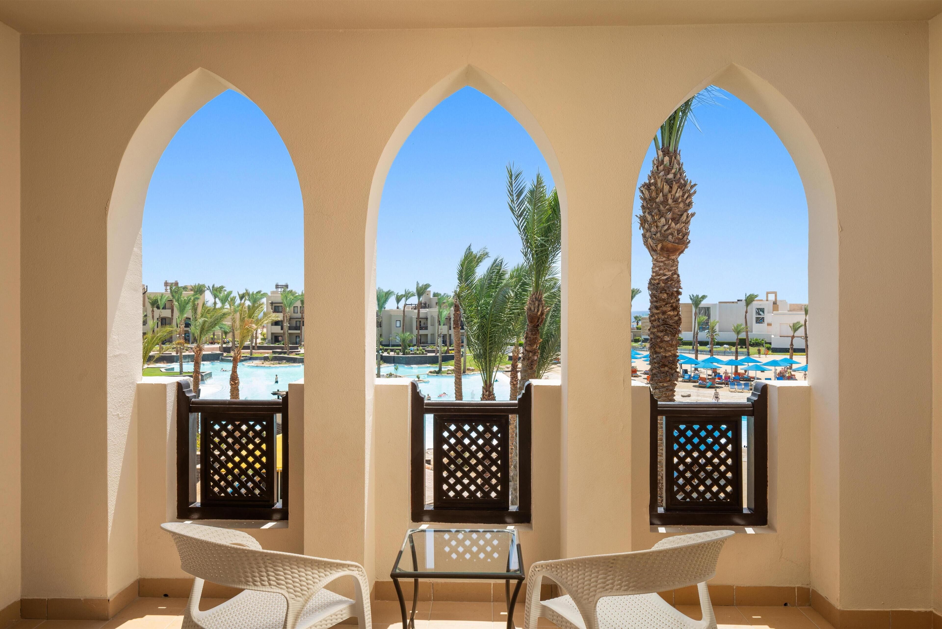 A balcony with three arched windows overlooking a pool area with palm trees and a clear blue sky.