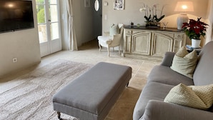 Sala de estar con un sofá gris, un reposapiés a juego, un sillón blanco y un mueble de madera. Hay un televisor de pantalla plana montado en la pared, una lámpara sobre una mesita auxiliar y una planta con flores rojas.