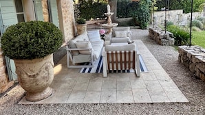Un patio exterior con una mesa de piedra, sillas blancas tipo lounge, una planta grande en maceta y una fuente de piedra.