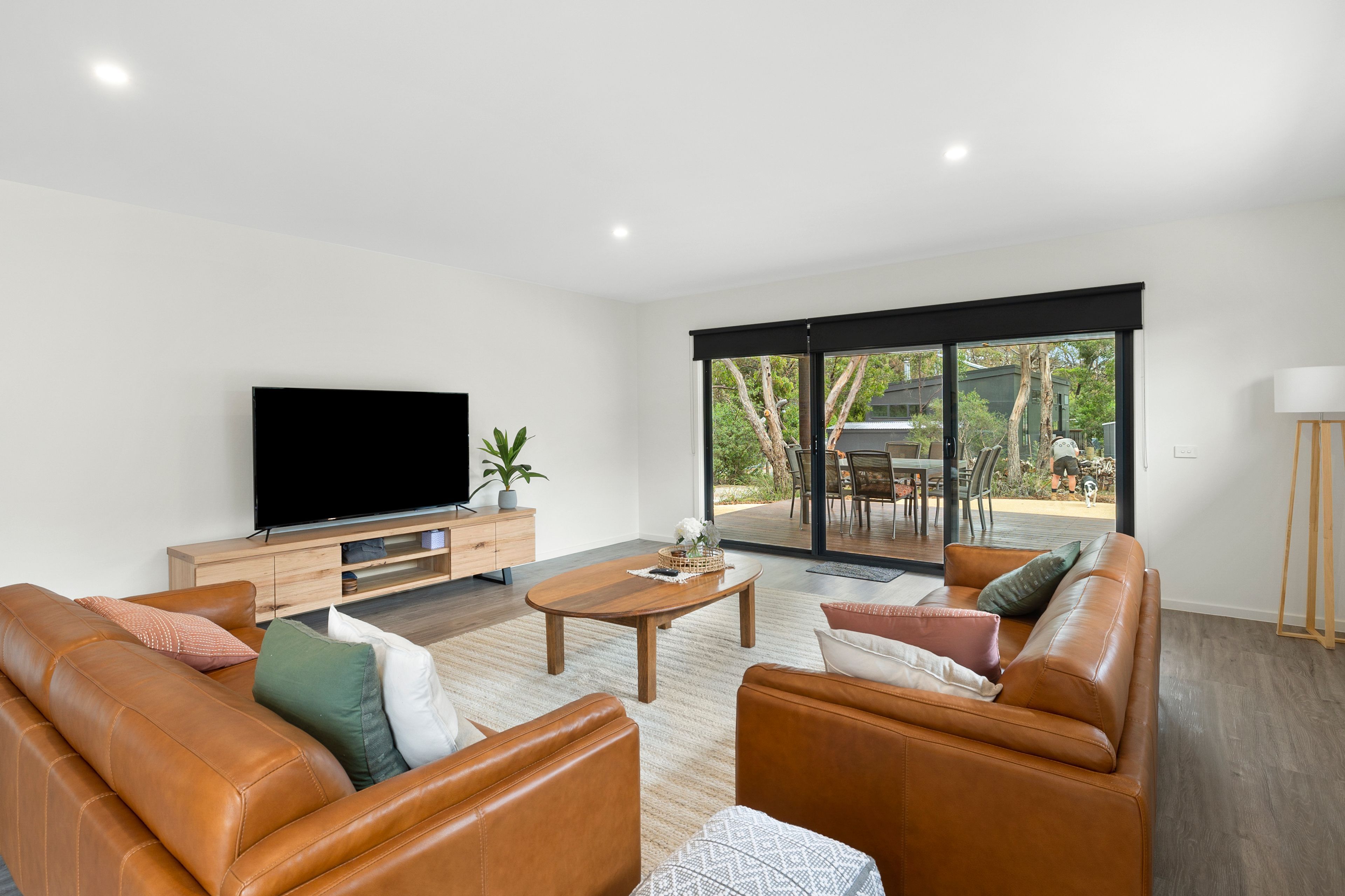Ein modernes Wohnzimmer mit Ledersofas, einem Flachbildfernseher, einem Holztisch und einer Glastür, die auf eine Terrasse im Freien führt.