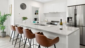 A modern kitchen with white cabinetry, a stainless steel refrigerator, a wine bottle, and a clock on the wall. - Chatley's (Aireys Inlet)