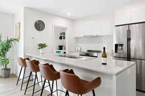 A modern kitchen with white cabinetry, a stainless steel refrigerator, a wine bottle, and a clock on the wall. - Chatley's (Aireys Inlet)