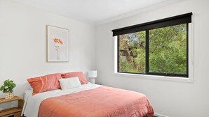 A bedroom with a bed, a window with a view of trees, a bedside table with a lamp, and a framed picture on the wall. - Chatley's (Aireys Inlet)