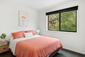 A bedroom with a bed, a window with a view of trees, a bedside table with a lamp, and a framed picture on the wall. - Chatley's (Aireys Inlet)