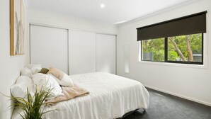 A bedroom with a bed, a window with a view of trees, and a plant on the floor. - Chatley's (Aireys Inlet)