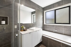 A modern bathroom with a glass shower enclosure, a white vanity, and a large window above the bathtub.