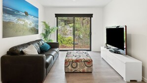 A modern living room with a leather sofa, a patterned ottoman, a flat-screen TV on a white stand, and a large painting of a beach scene. - Chatley's (Aireys Inlet)