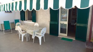 Un espace extérieur aménagé pour les repas, avec une table et des chaises, des volets verts et un sol carrelé.