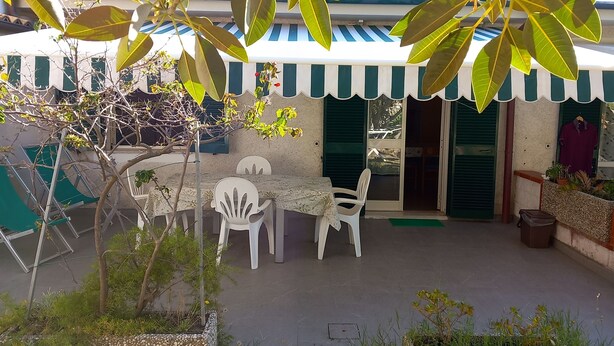Une terrasse extérieure avec une table et des chaises, entourée de verdure et bordée d’un bâtiment aux volets verts.